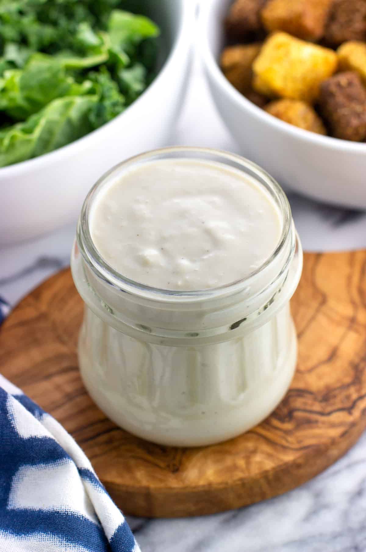 A glass jar of caesar dressing with salad ingredients in the background.
