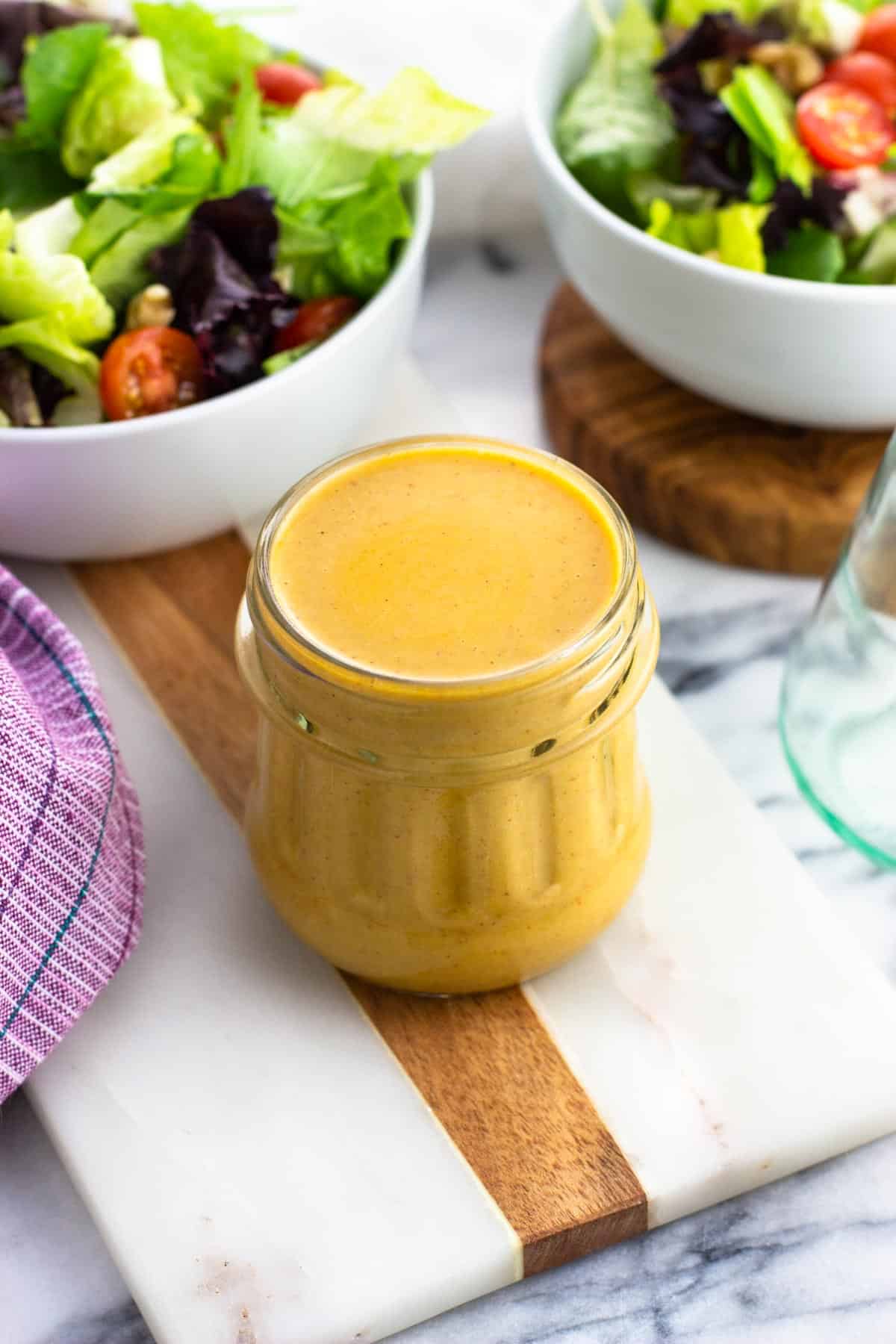 A jar of hummus salad dressing surrounded by bowls of salad.