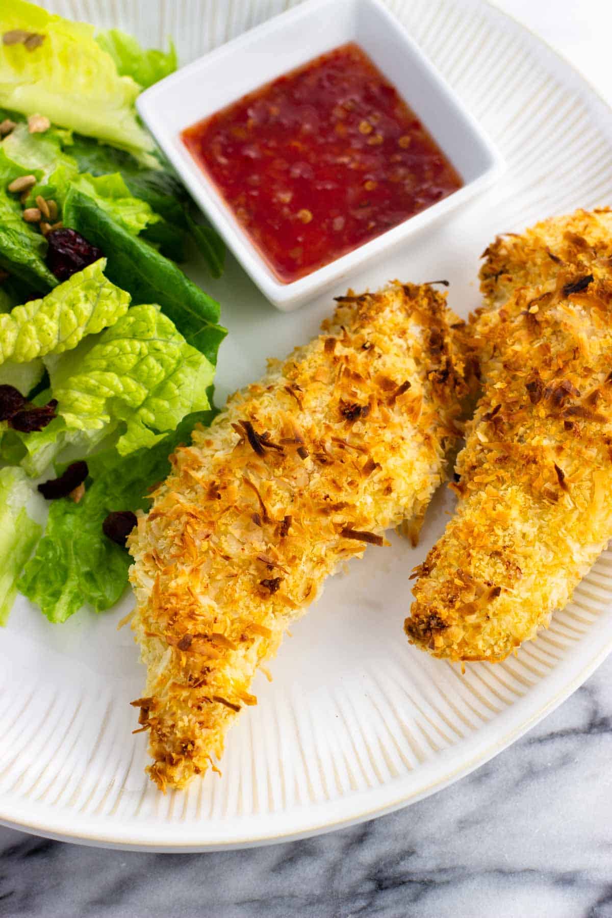 Two chicken tenders on a plate with salad and dipping sauce.