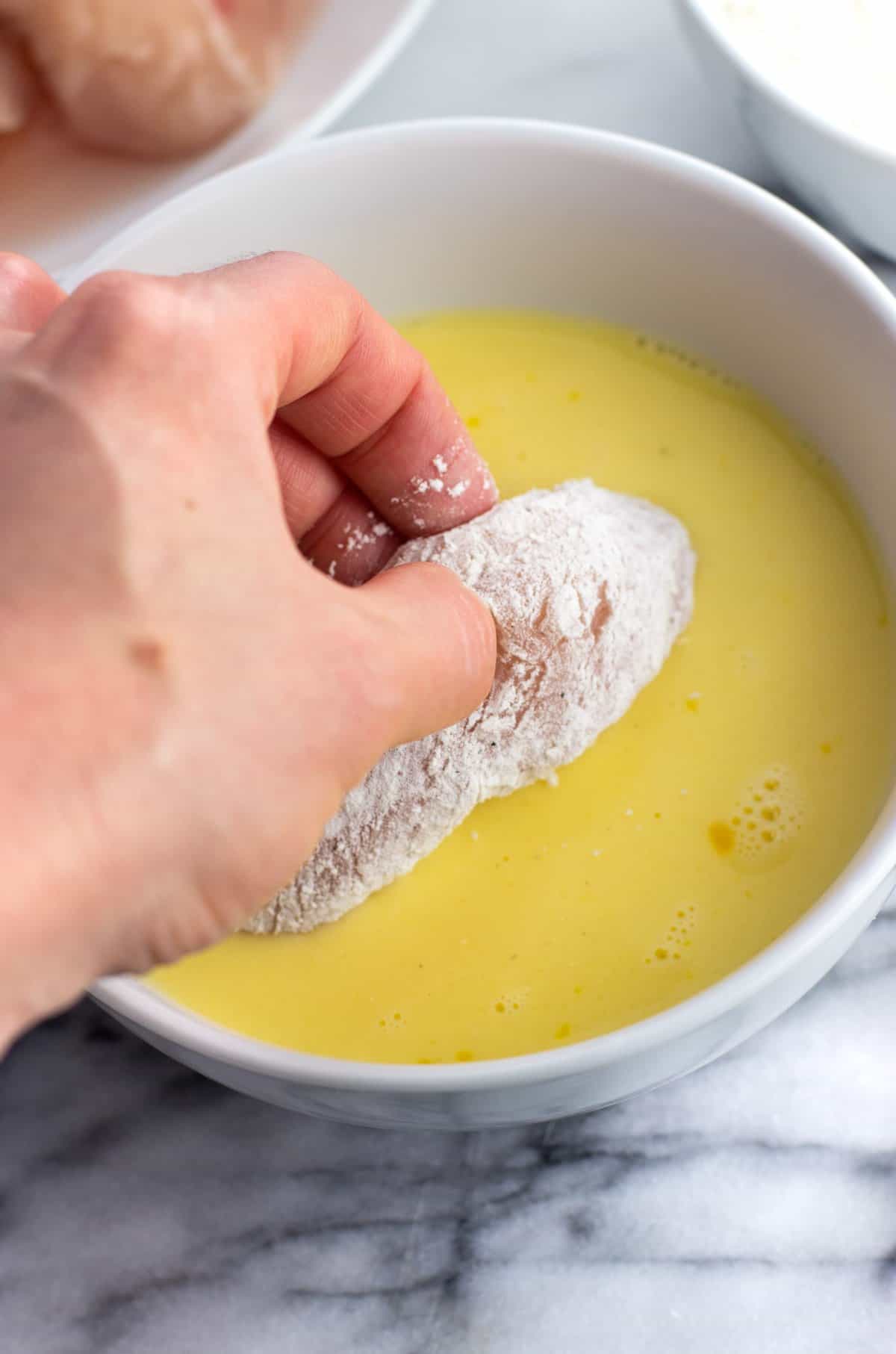 A hand dipping a flour dredged tender in egg.