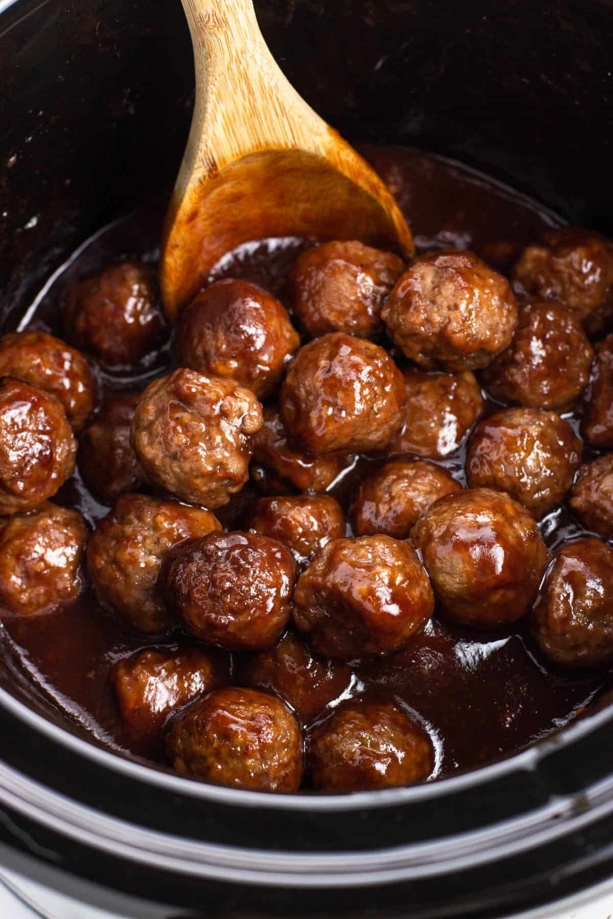 Sauce-coated meatballs in a slow cooker with a wooden spoon.