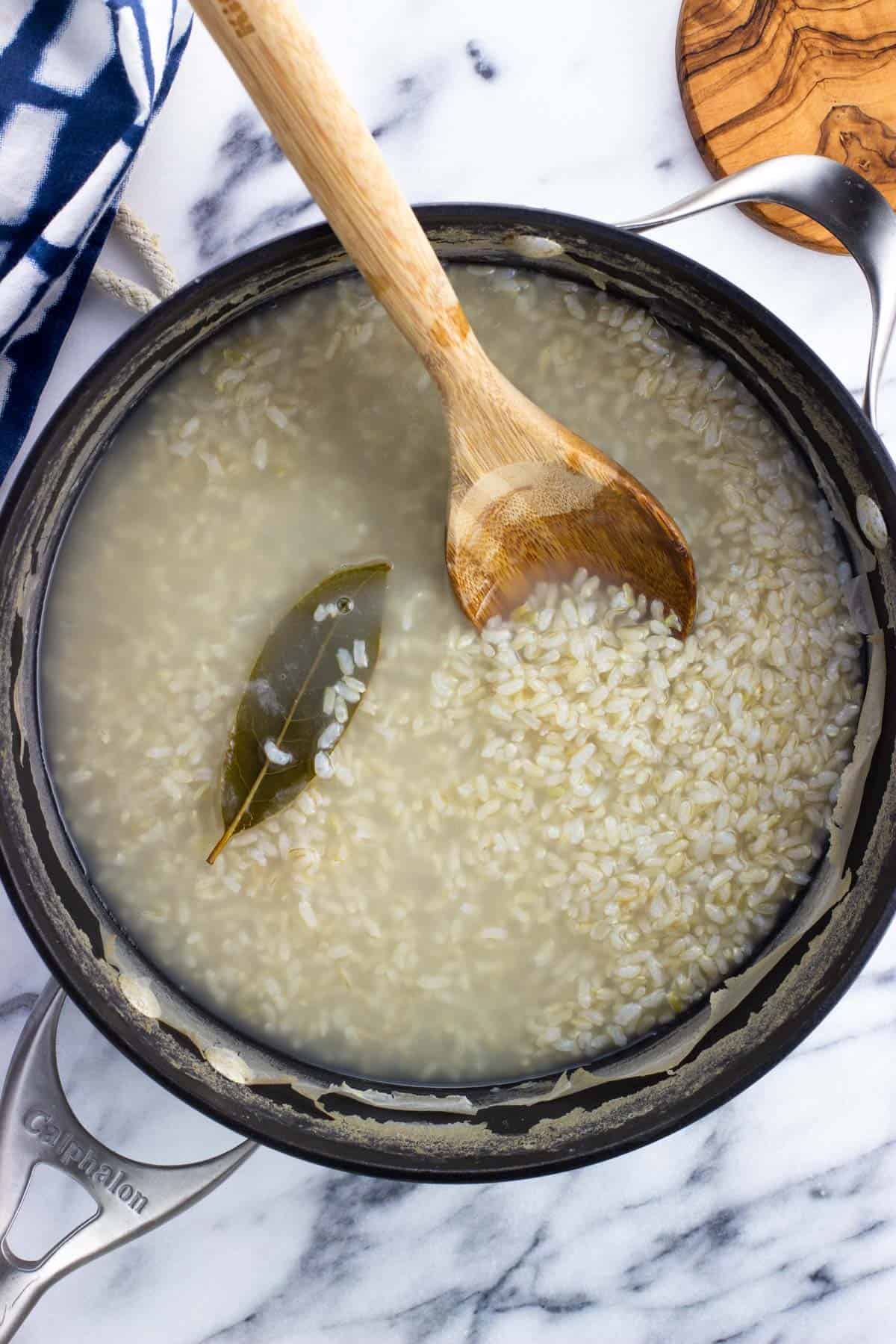 Rice, water, and a bay leaf in a pan with a wooden spoon.