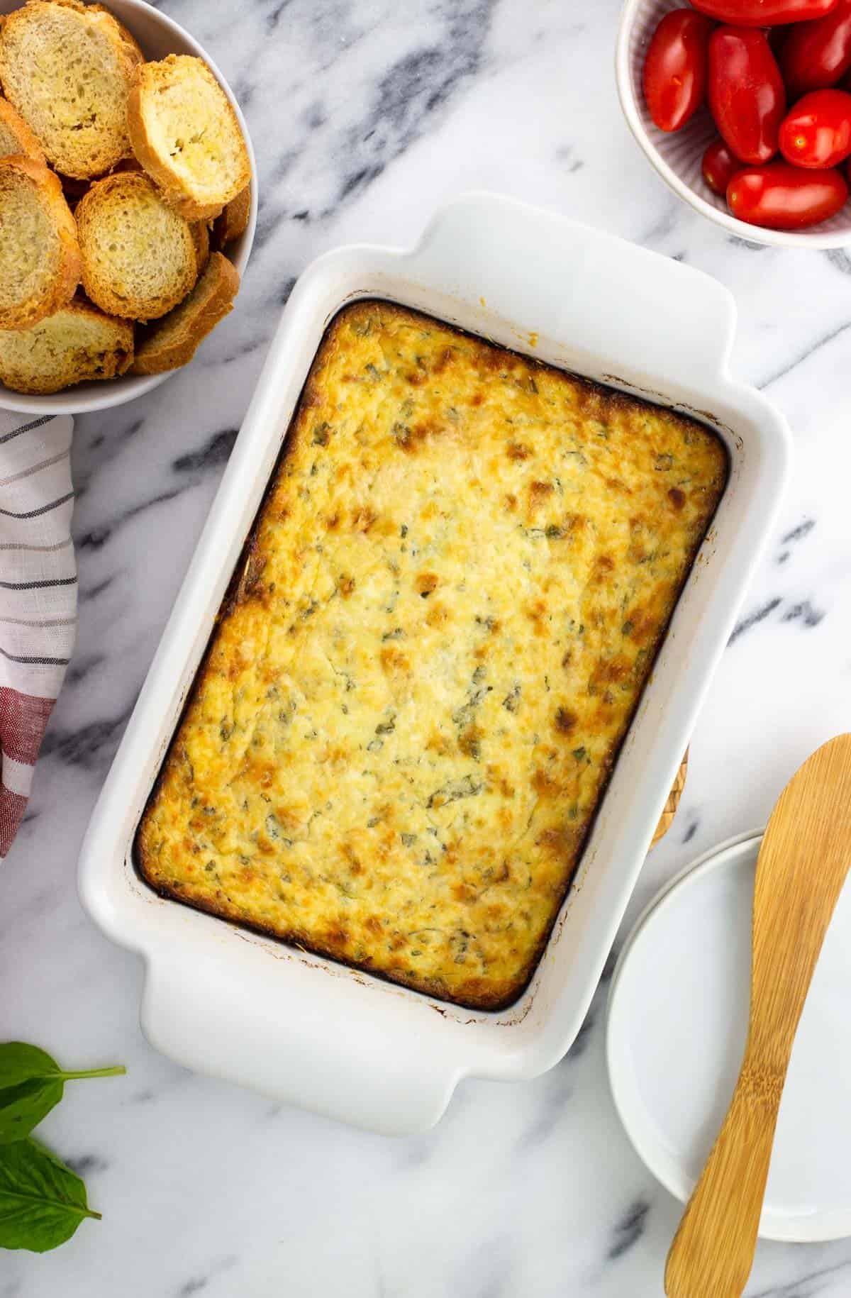 A rectangular dish of baked ricotta surrounded by crostini and tomatoes.