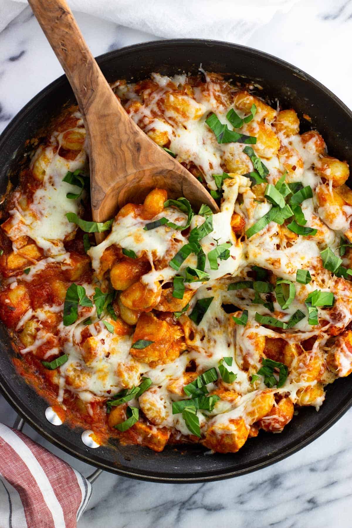 A skillet of chicken Parmesan gnocchi with a wooden serving spoon in it.