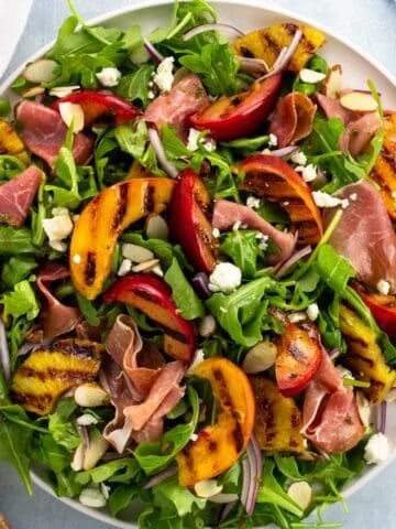 A large plate topped with arugula, grilled fruits, feta, almonds, red onion, and vinaigrette.