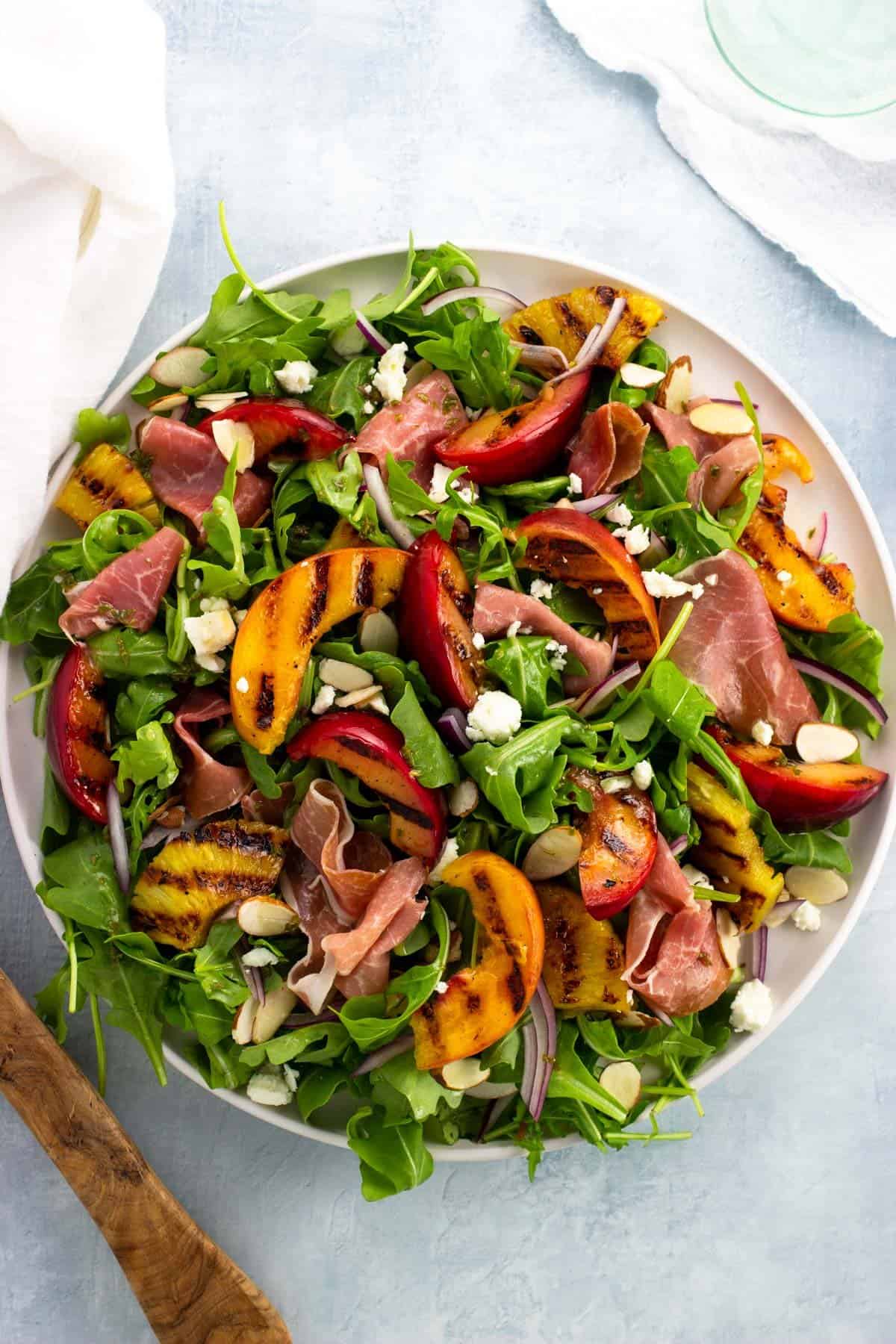 A large plate topped with arugula, grilled fruits, prosciutto, feta, almonds, red onion, and vinaigrette.