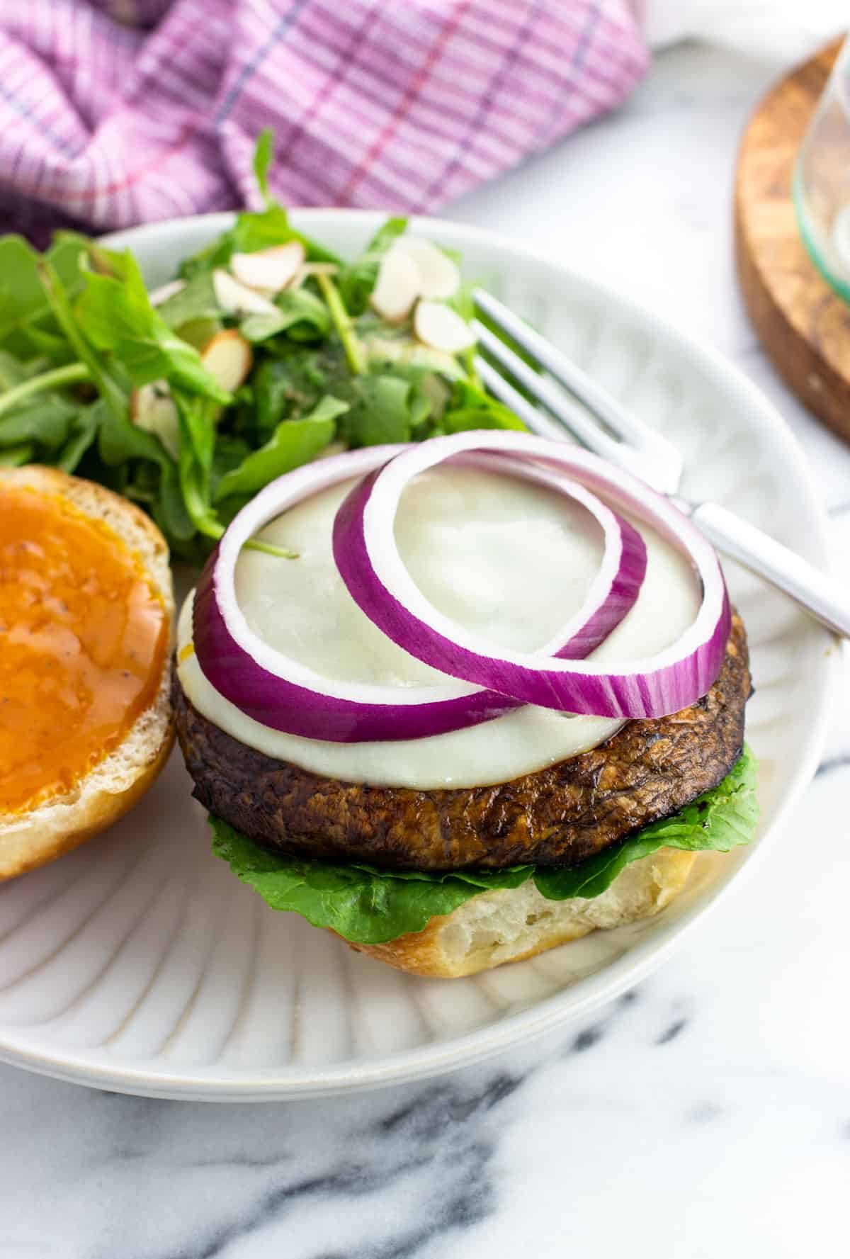 An open-face portobello mushroom burger topped with cheese and red onion on a plate with a salad.