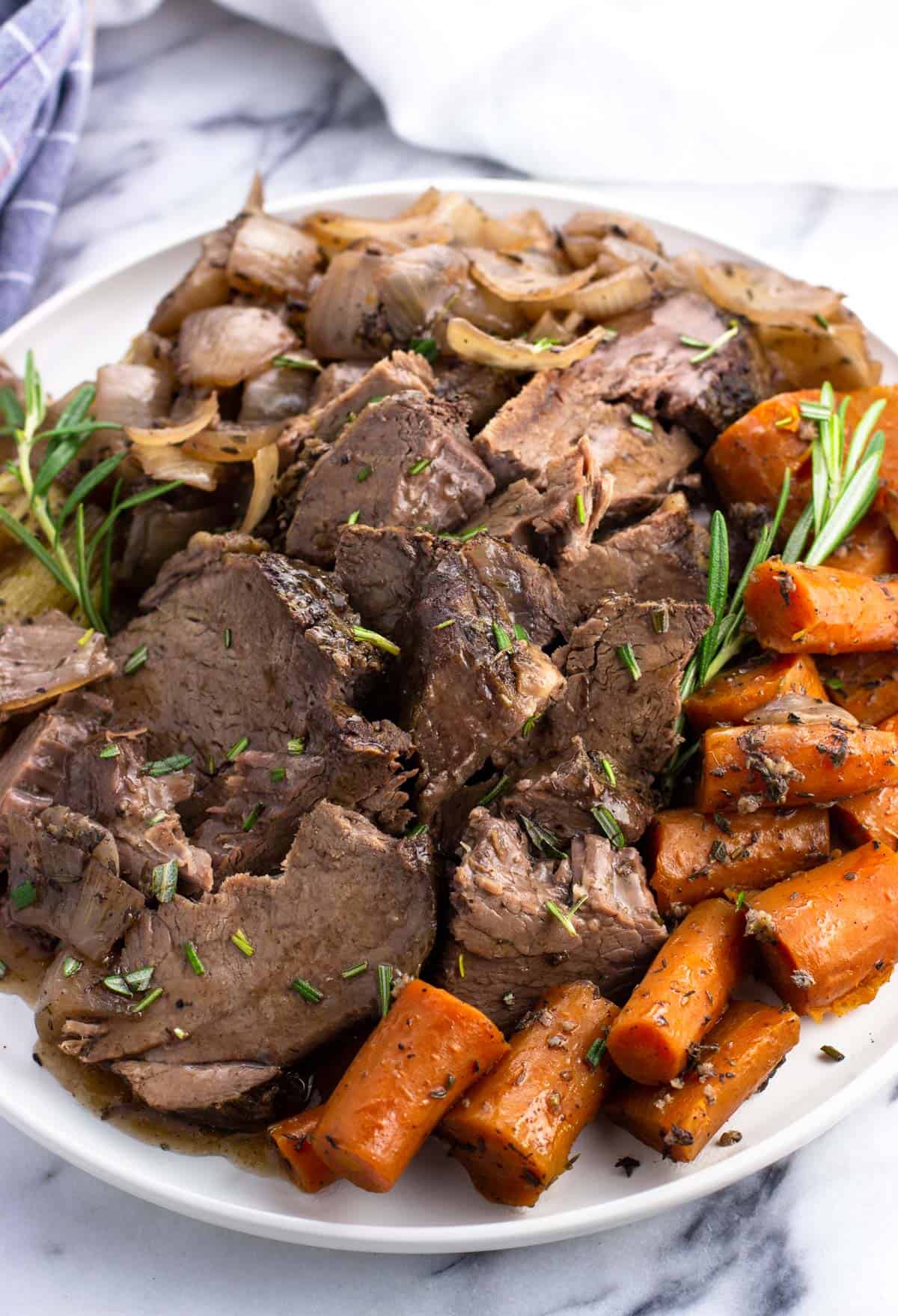 Sliced beef shoulder roast, carrots, celery, onions, and rosemary sprigs on a plate.