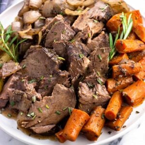 Sliced beef shoulder roast, carrots, celery, onions, and rosemary sprigs on a plate.