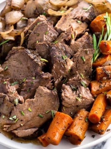 Sliced beef shoulder roast, carrots, celery, onions, and rosemary sprigs on a plate.