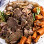 Sliced beef shoulder roast, carrots, celery, onions, and rosemary sprigs on a plate.