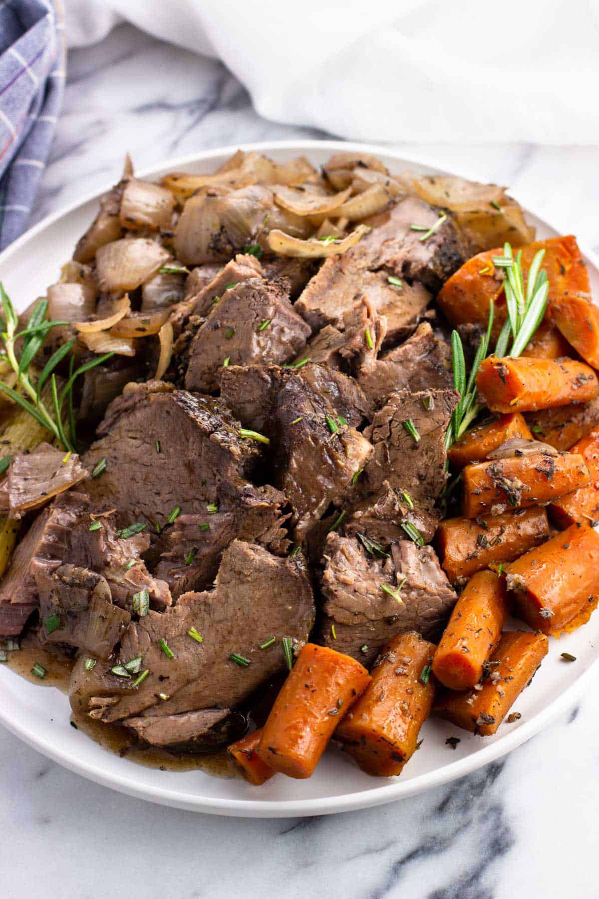 Sliced beef shoulder roast, carrots, celery, onions, and rosemary sprigs on a plate.
