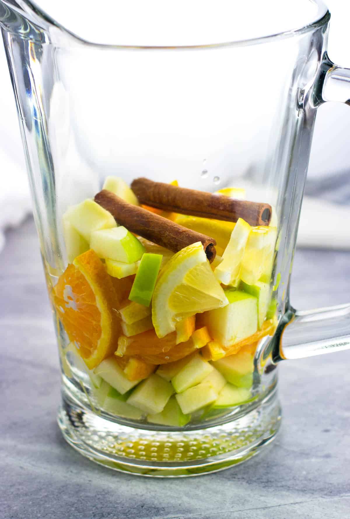 Fruit and two cinnamon sticks in the bottom of a glass pitcher.