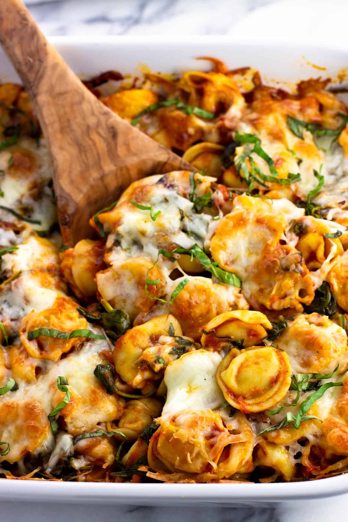 A wooden spoon scooping out a serving of baked tortellini from the dish.