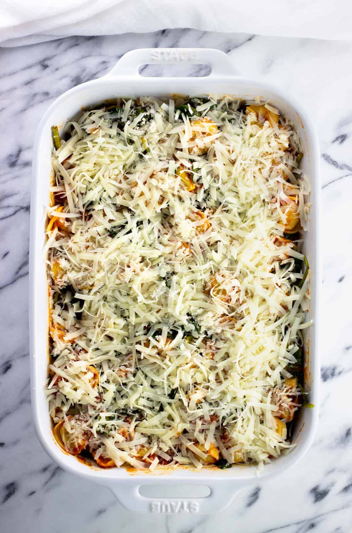 Tortellini, sauce, and spinach added to a baking dish topped with shredded cheeses.