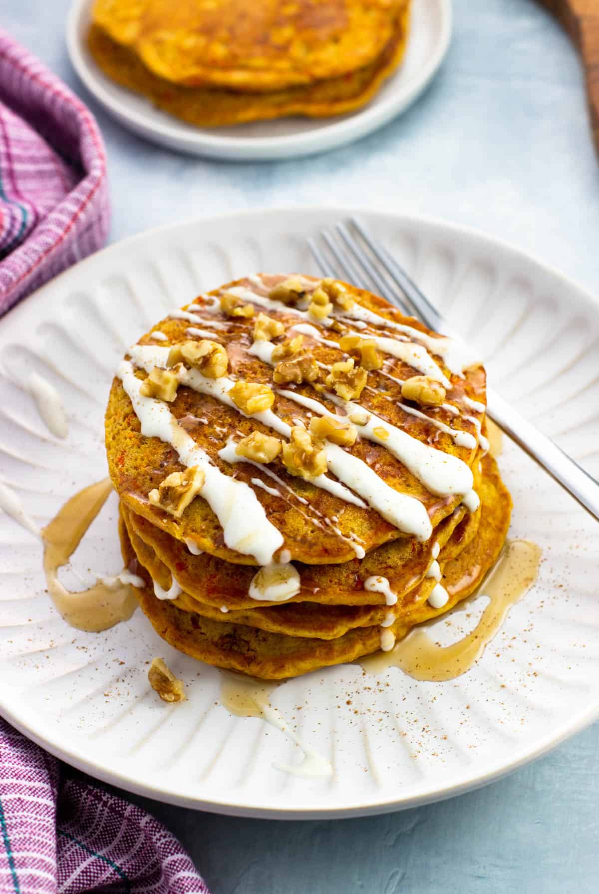 A stack of carrot cake pancakes served with cream cheese glaze, maple syrup, and walnuts.