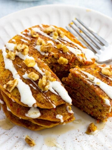 A stack of cream cheese and maple syrup drizzled carrot cake pancakes with a wedge cut out.