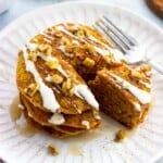 A stack of cream cheese and maple syrup drizzled carrot cake pancakes with a wedge cut out.