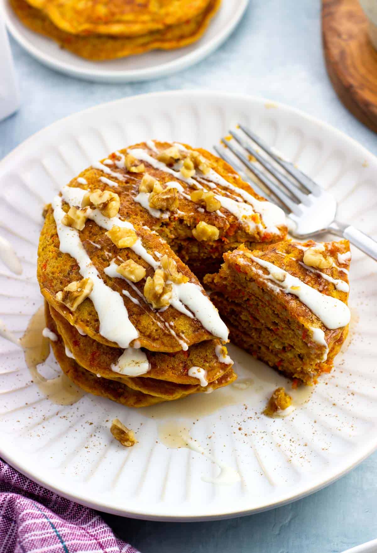 A stack of cream cheese and maple syrup drizzled carrot cake pancakes with a wedge cut out.