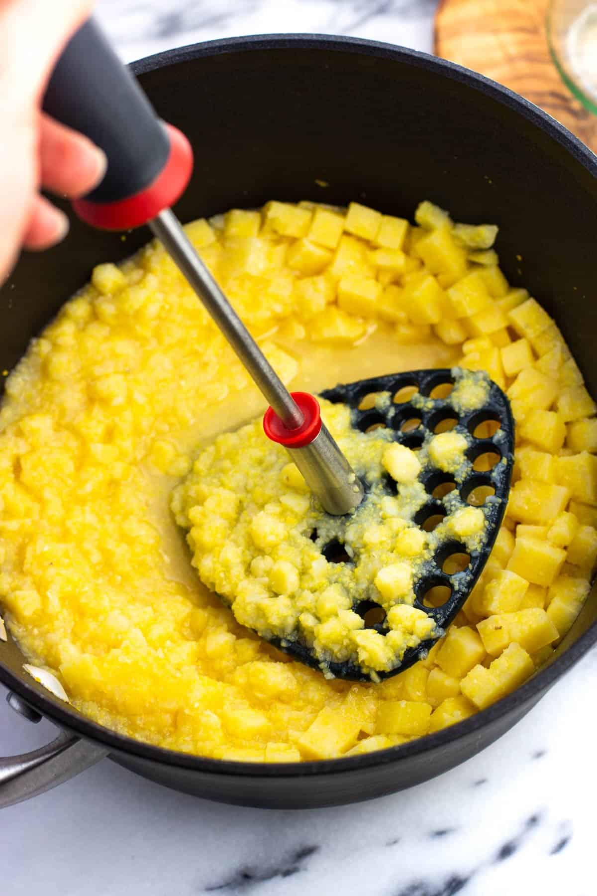 A potato masher mashing the cubes of warm cubes of polenta in broth.