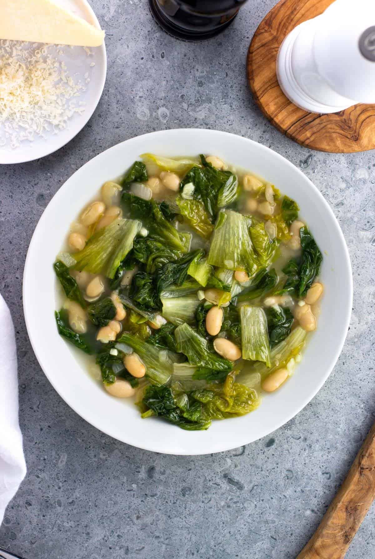 A serving of escarole and beans in a bowl surrounded by a grated wedge of Parmesan and salt and pepper mills.