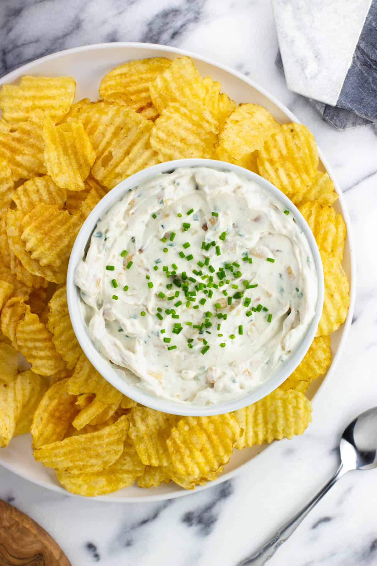 A bowl of sour cream and onion dip on a plate surrounded by ruffle chips.