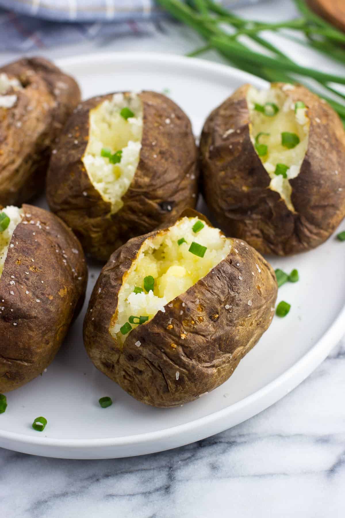 Five baked potatoes on a plate open with butter and chives.