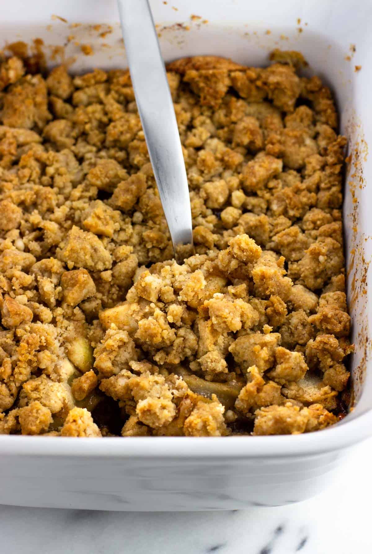 A pan of apple crumble with a serving spoon scooping a portion.