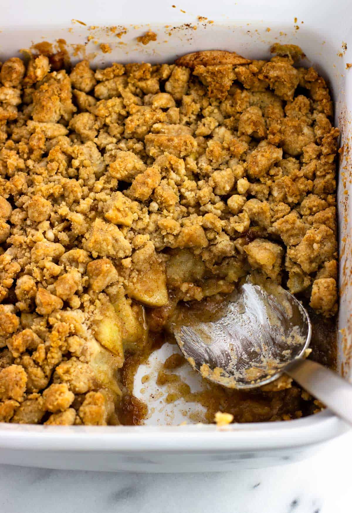 A baking dish of apple crumble with a serving spoon with a portion removed to show the layers.