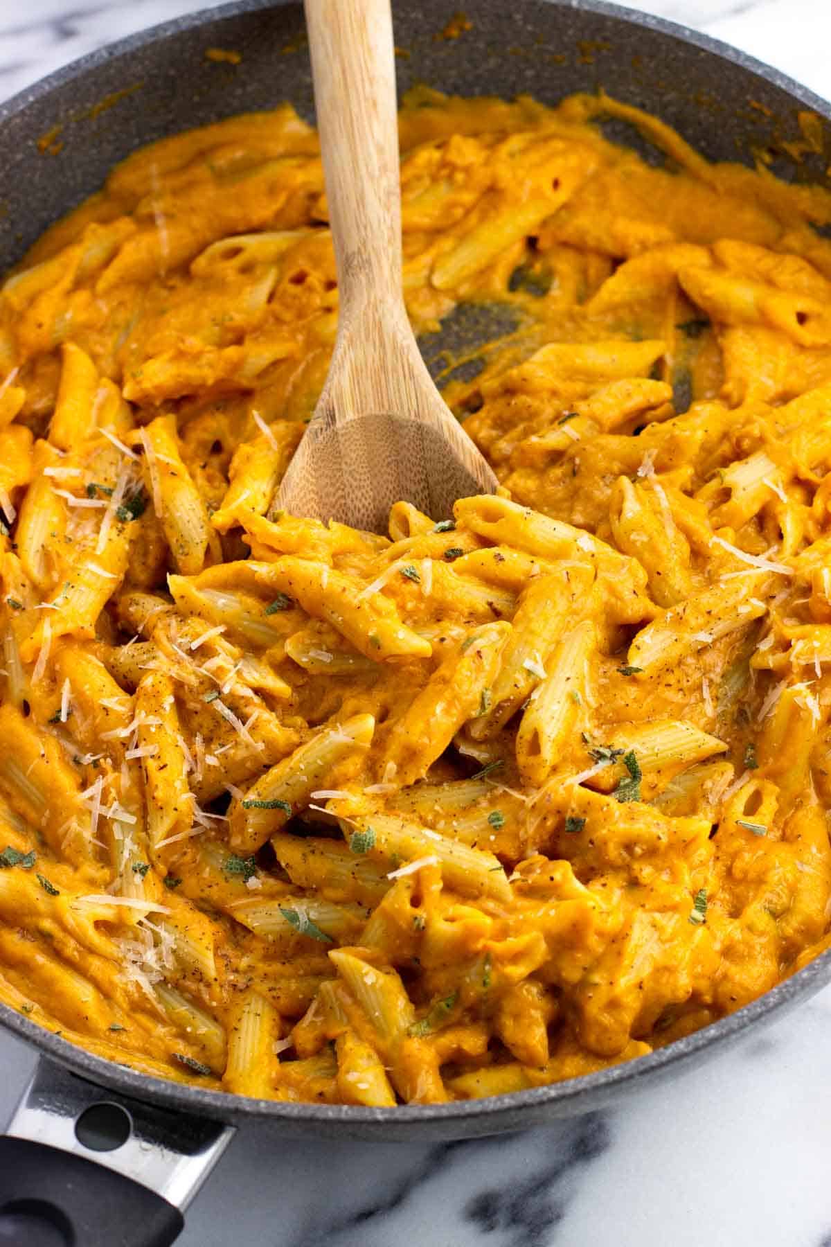 A skillet of penne pasta tossed in pumpkin sauce with a wooden serving spoon.