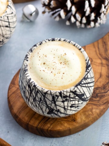 An eggnog latte in a ceramic glass surrounded by pine cones and jingle bells.