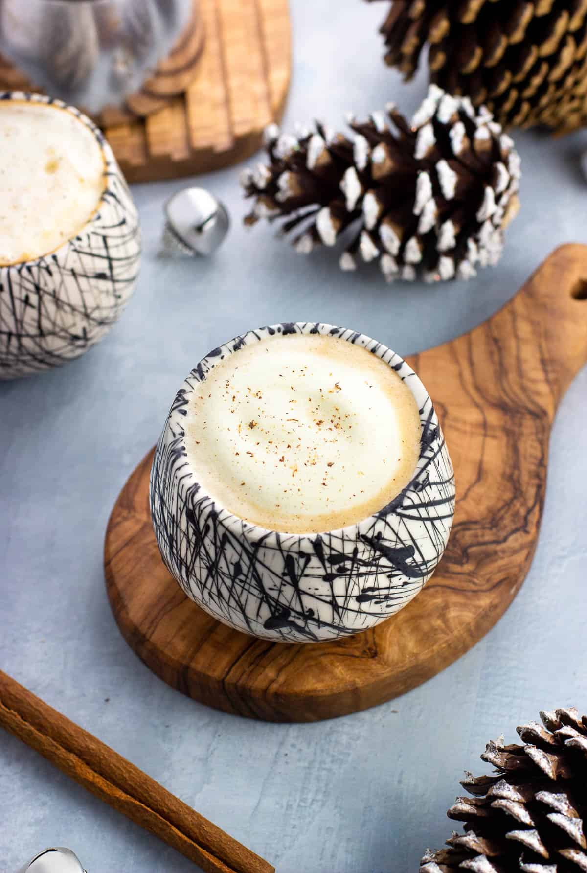 An eggnog latte in a ceramic glass surrounded by pine cones and jingle bells.