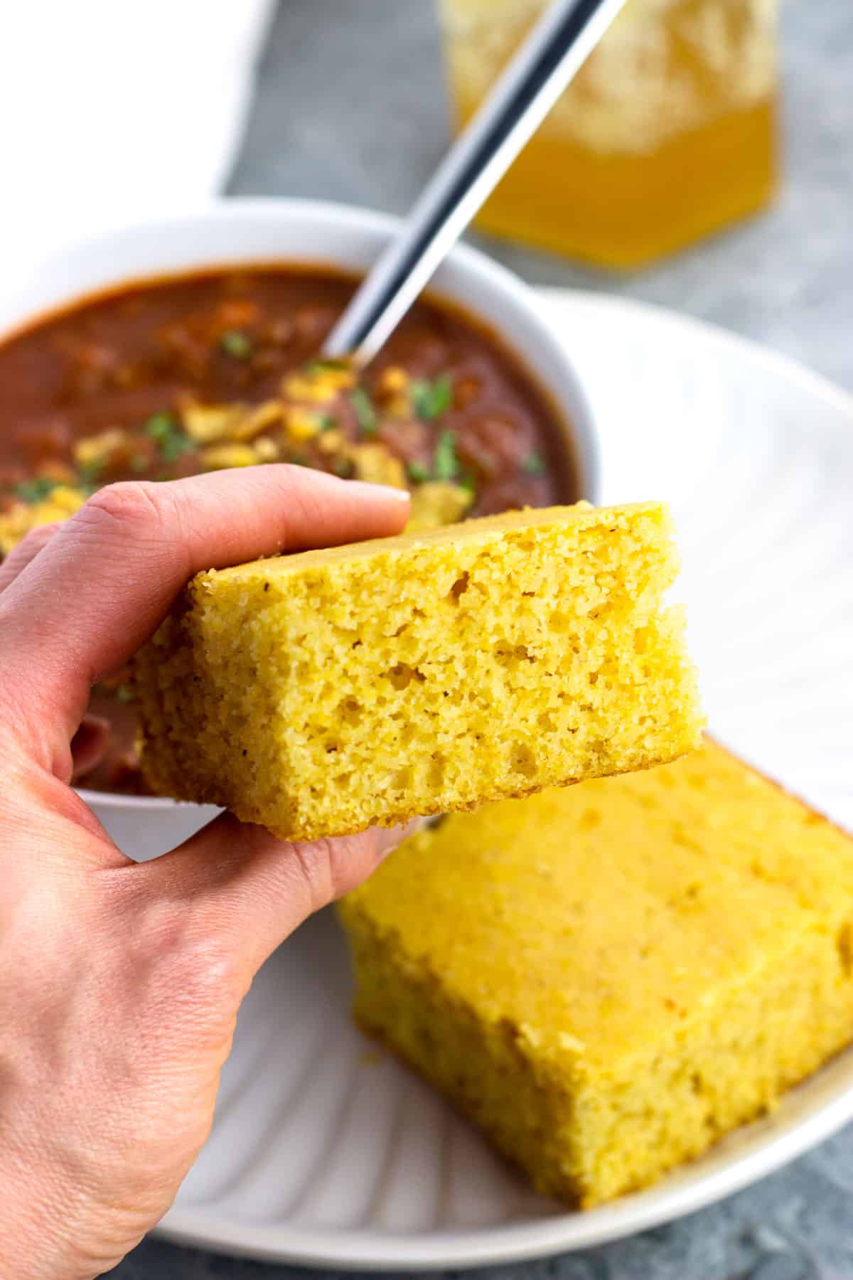 A hand holding a square of cornbread in front of a bowl of chili.