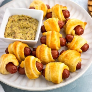 A plate of pigs in a blanket with a small bowl of dipping mustard.