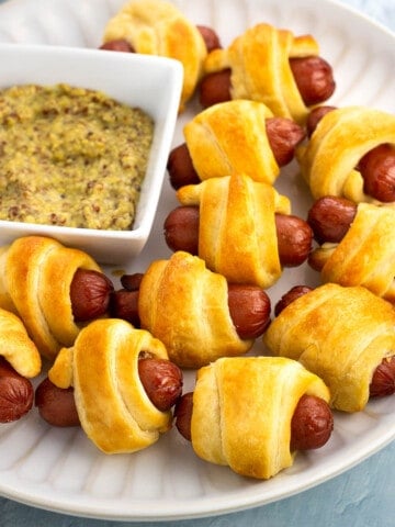 A plate of pigs in a blanket with a small bowl of dipping mustard.