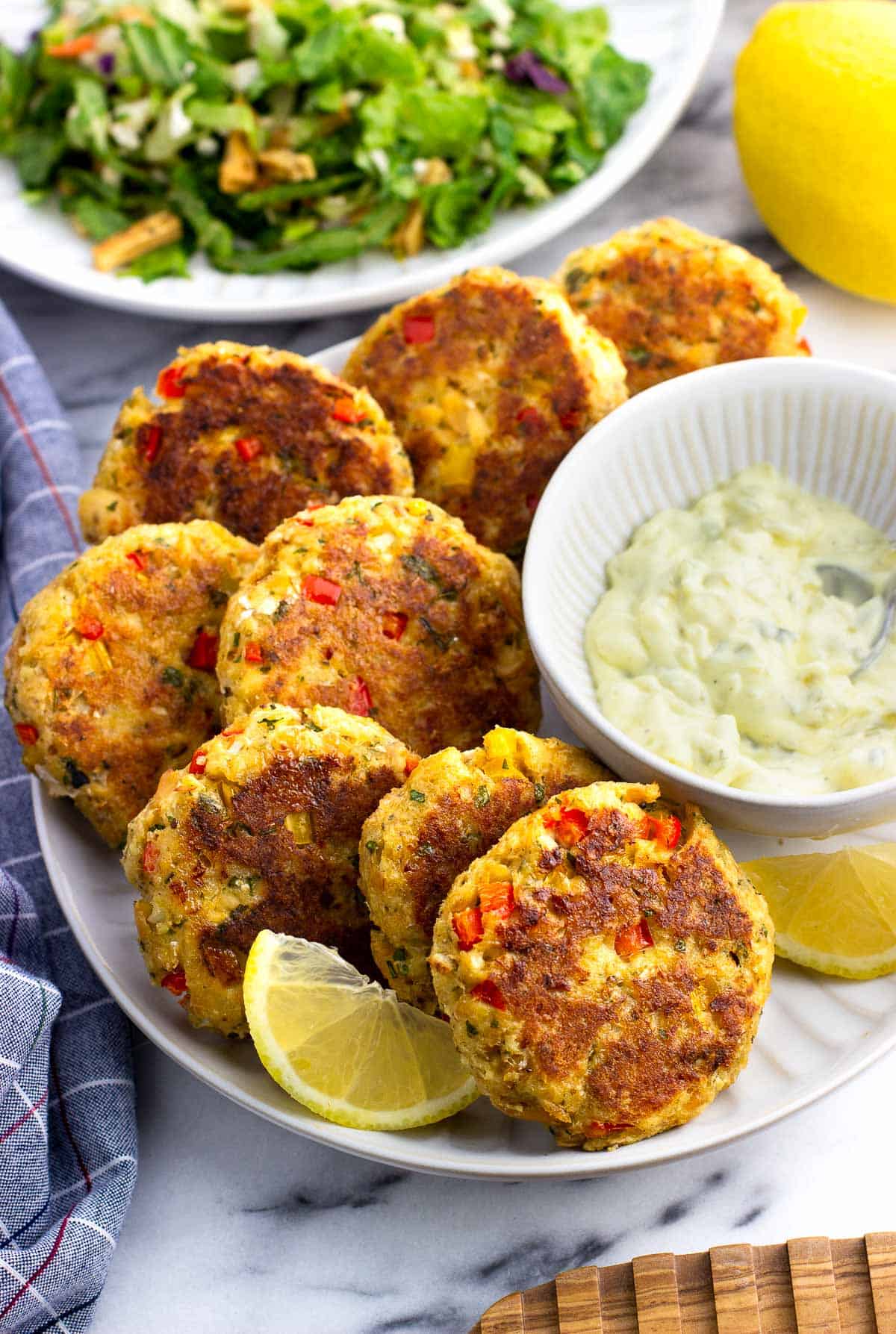 Baked salmon patties on a plate with tartar sauce and lemon wedges in front of a plate of salad.