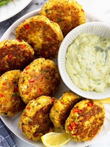 Eight salmon patties on a plate with a bowl of tartar sauce and lemon wedges.