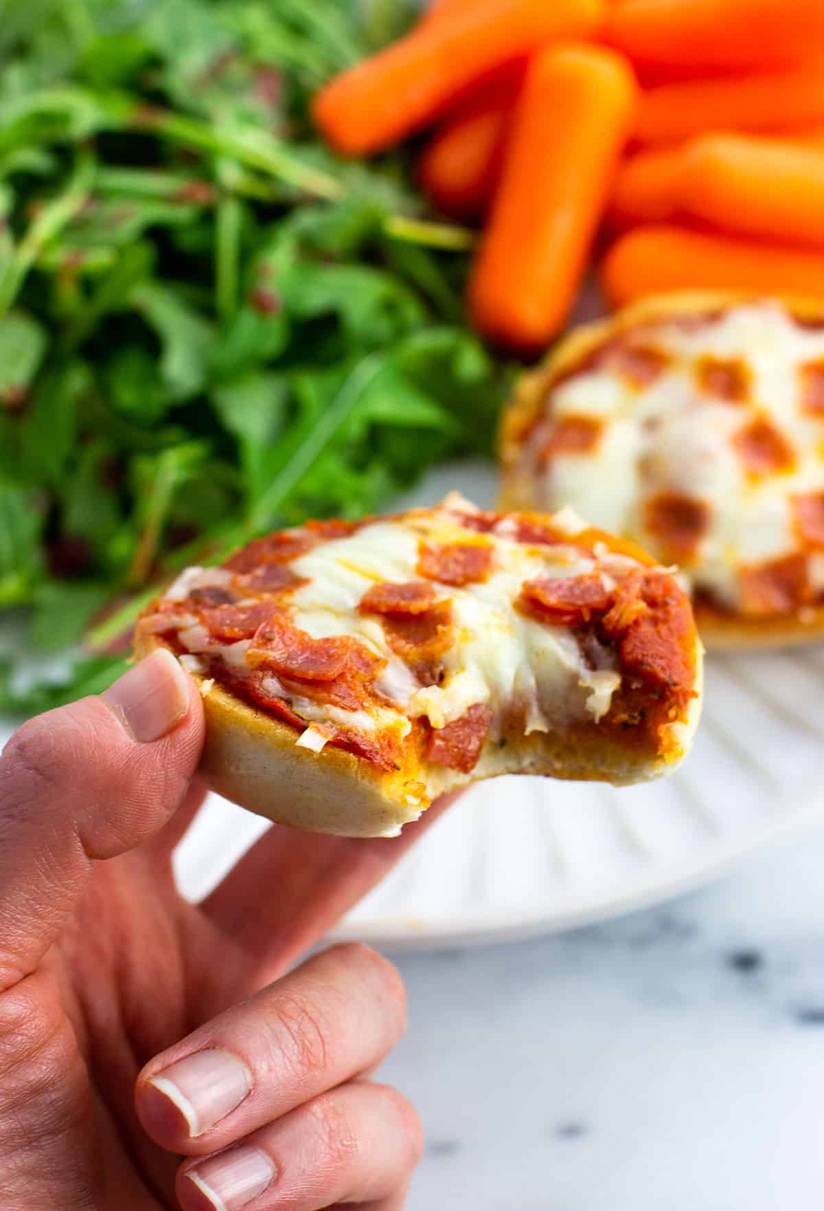 A hand holding up a mini pizza bagel in front of a plate of salad and carrots.