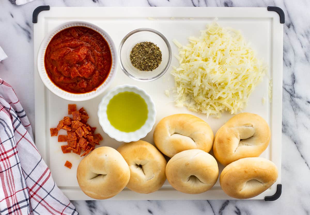 Pizza bagel ingredients on a cutting board in separate containers.