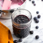 A jar of blueberry maple syrup surrounded by whole blueberries.