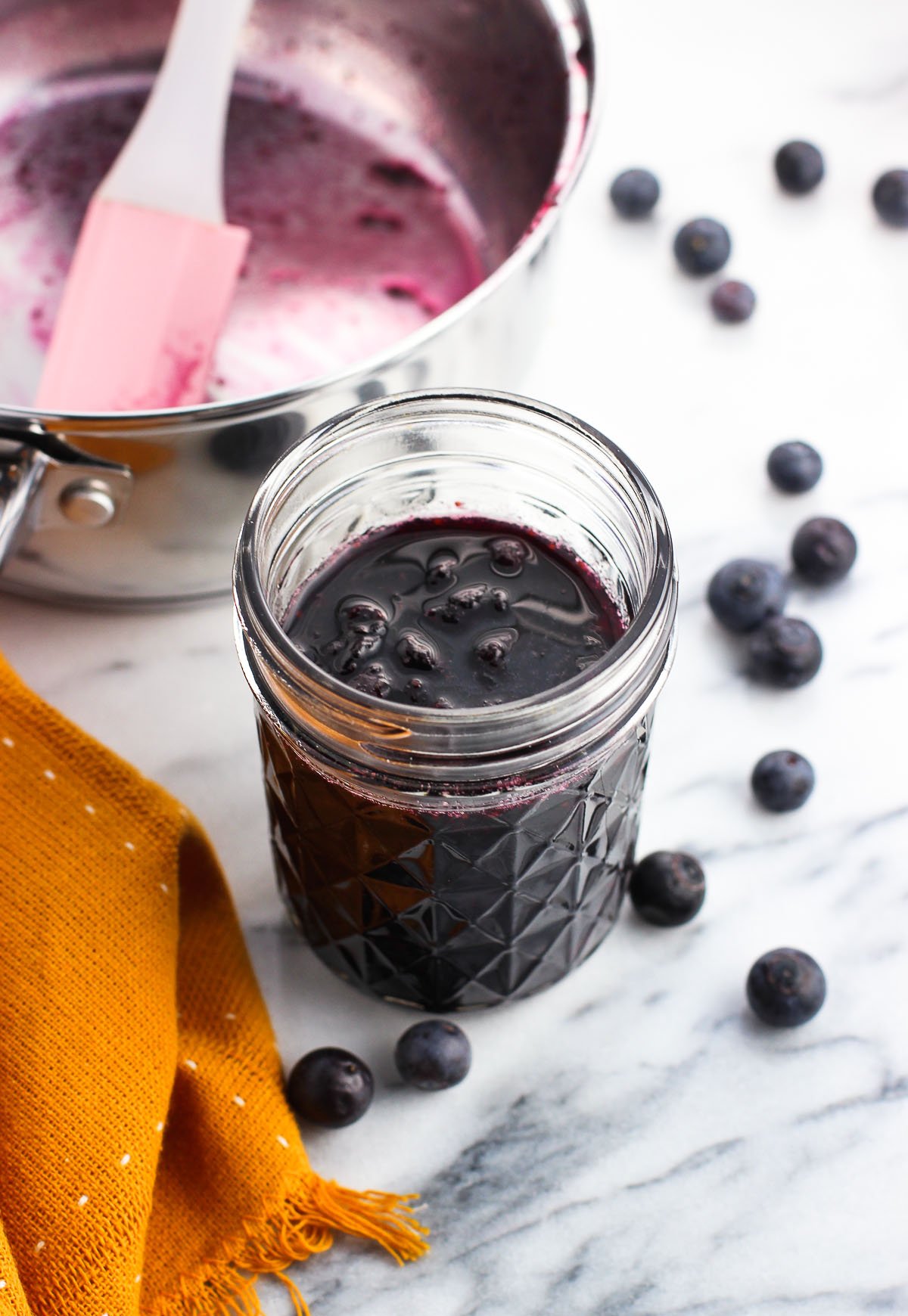 A jar of blueberry maple syrup surrounded by whole blueberries.