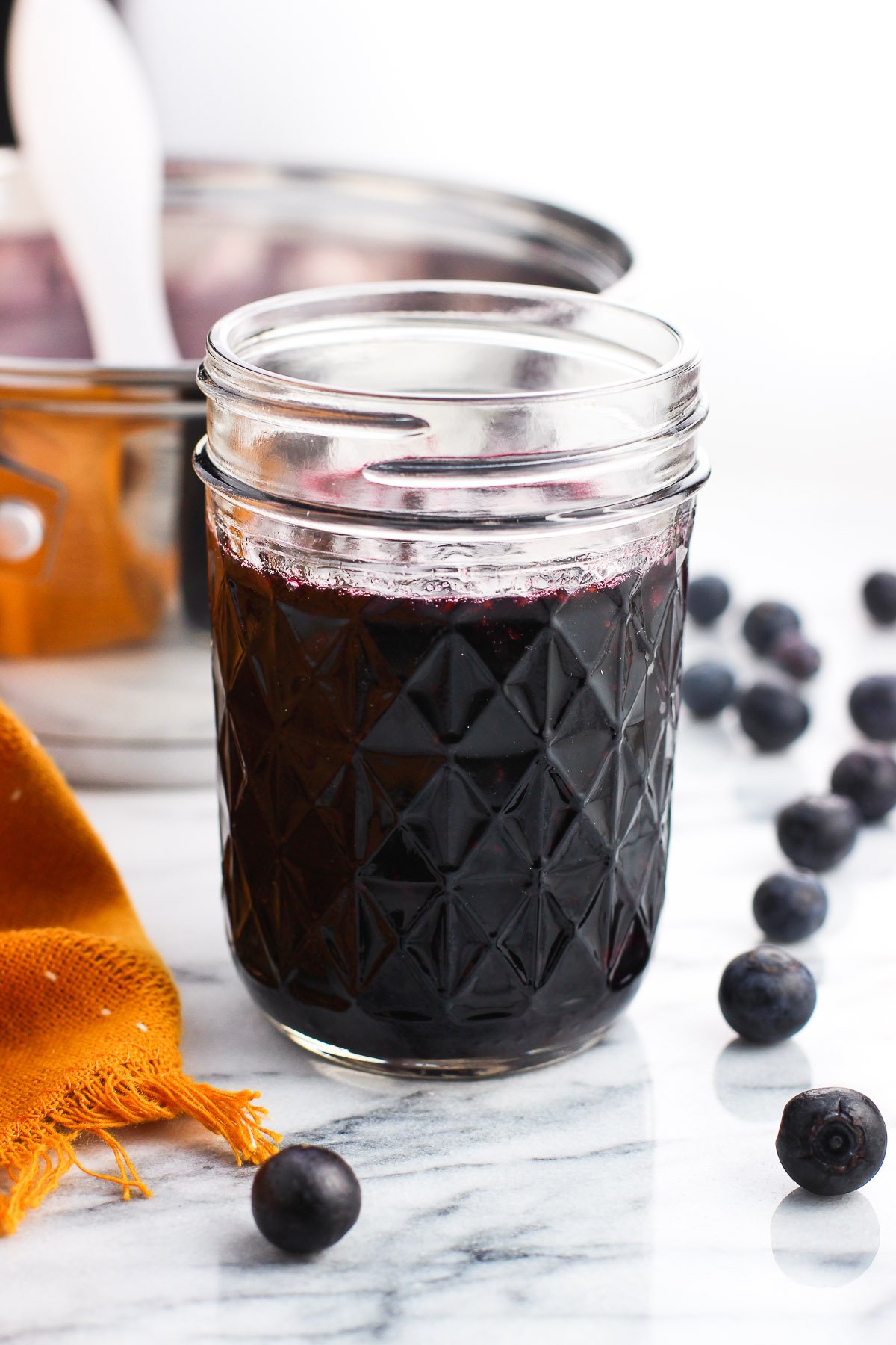 A jar of blueberry maple syrup surrounded by whole blueberries.