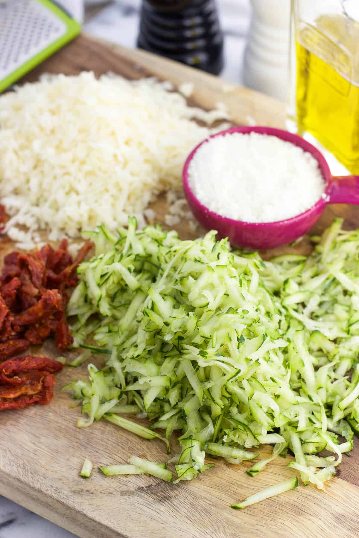Shredded cheese, zucchini, and sun-dried tomatoes on a wooden cutting board.