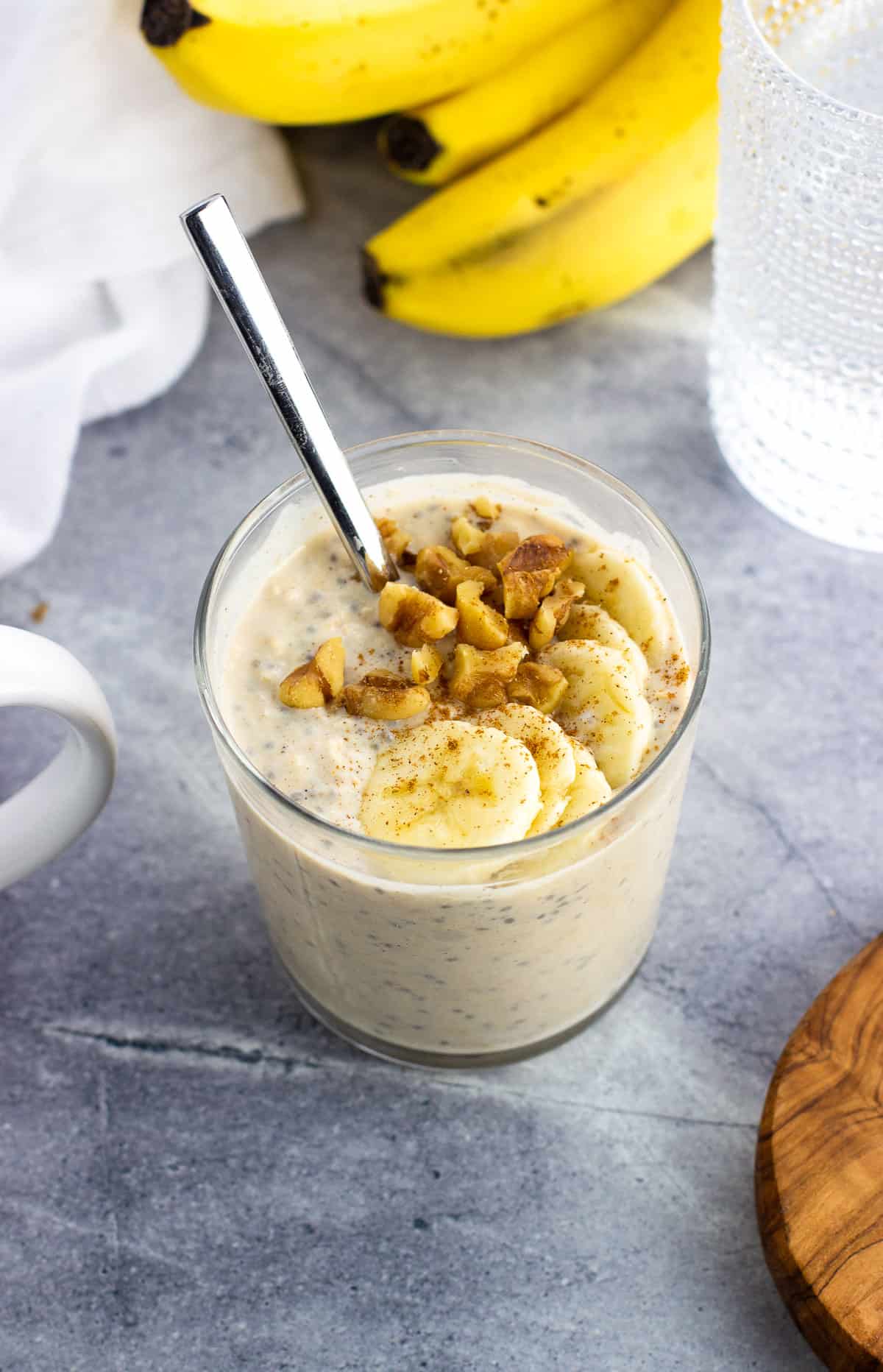 A short glass of creamy overnight oats topped with walnuts and banana slices with a spoon in it.