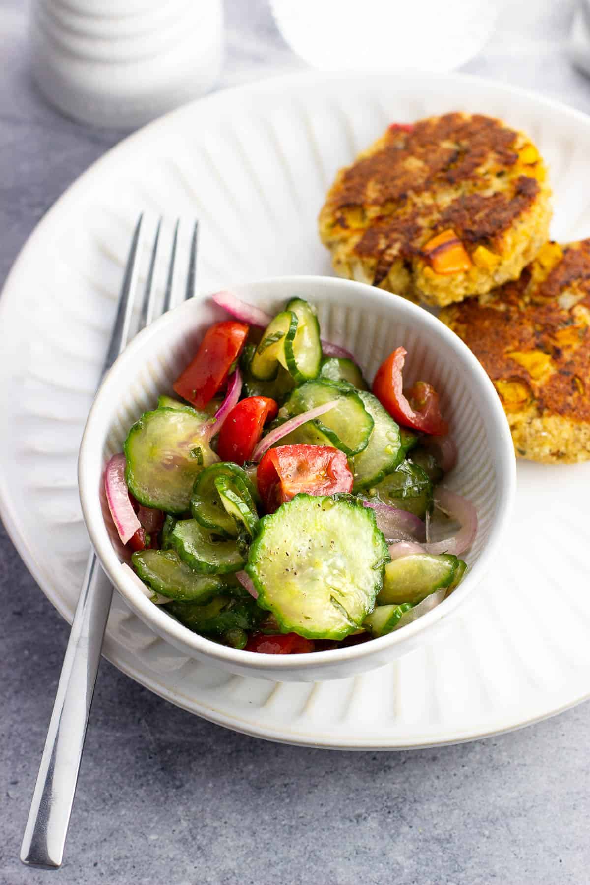 A small side-dish bowl of marinated cucumber salad on a plate next to salmon patties.