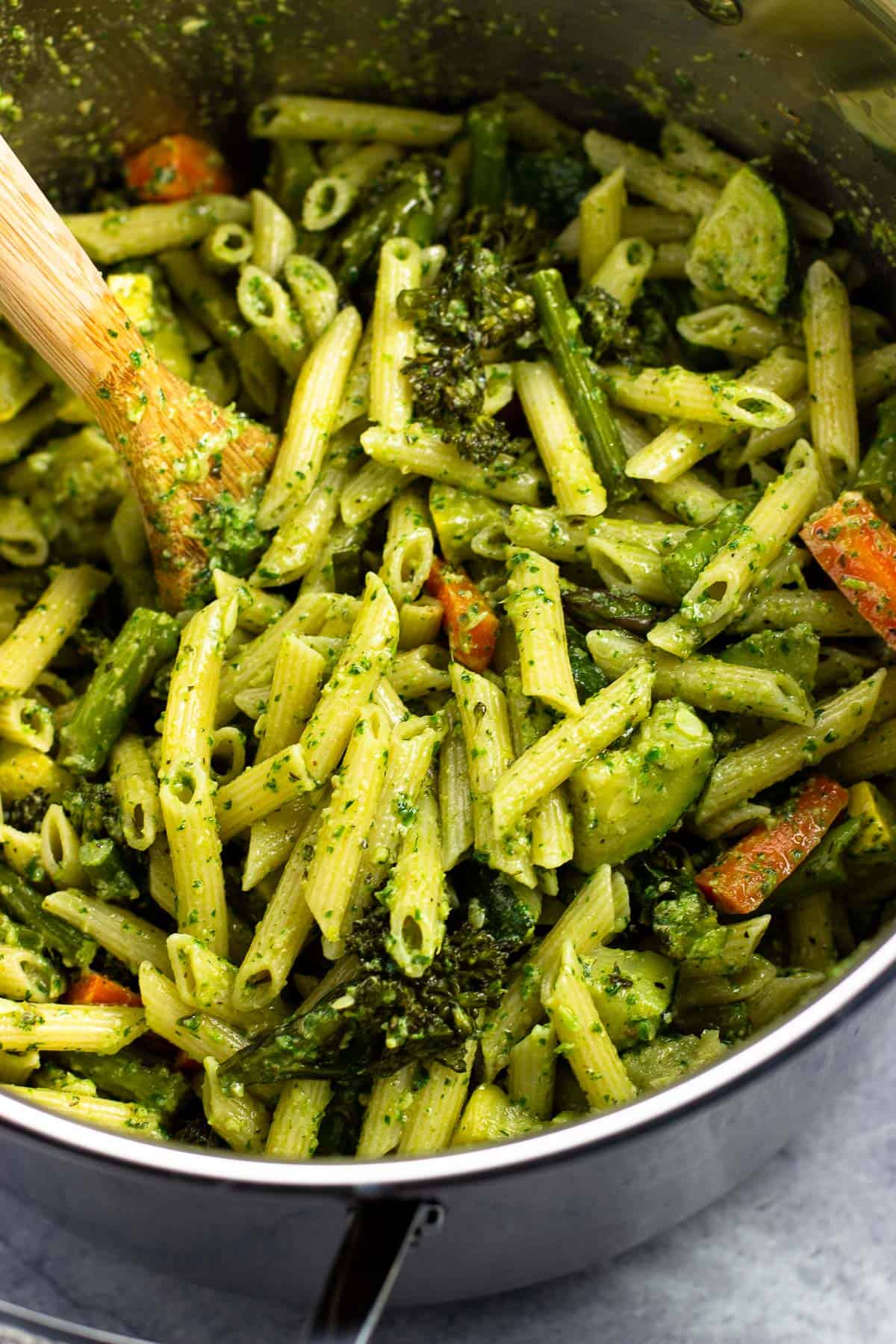 Pesto pasta primavera stirred together in a pot with a wooden spoon.