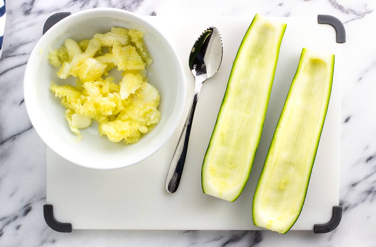 A halved zucchini on a cutting board with the middles scooped out and put into a bowl.