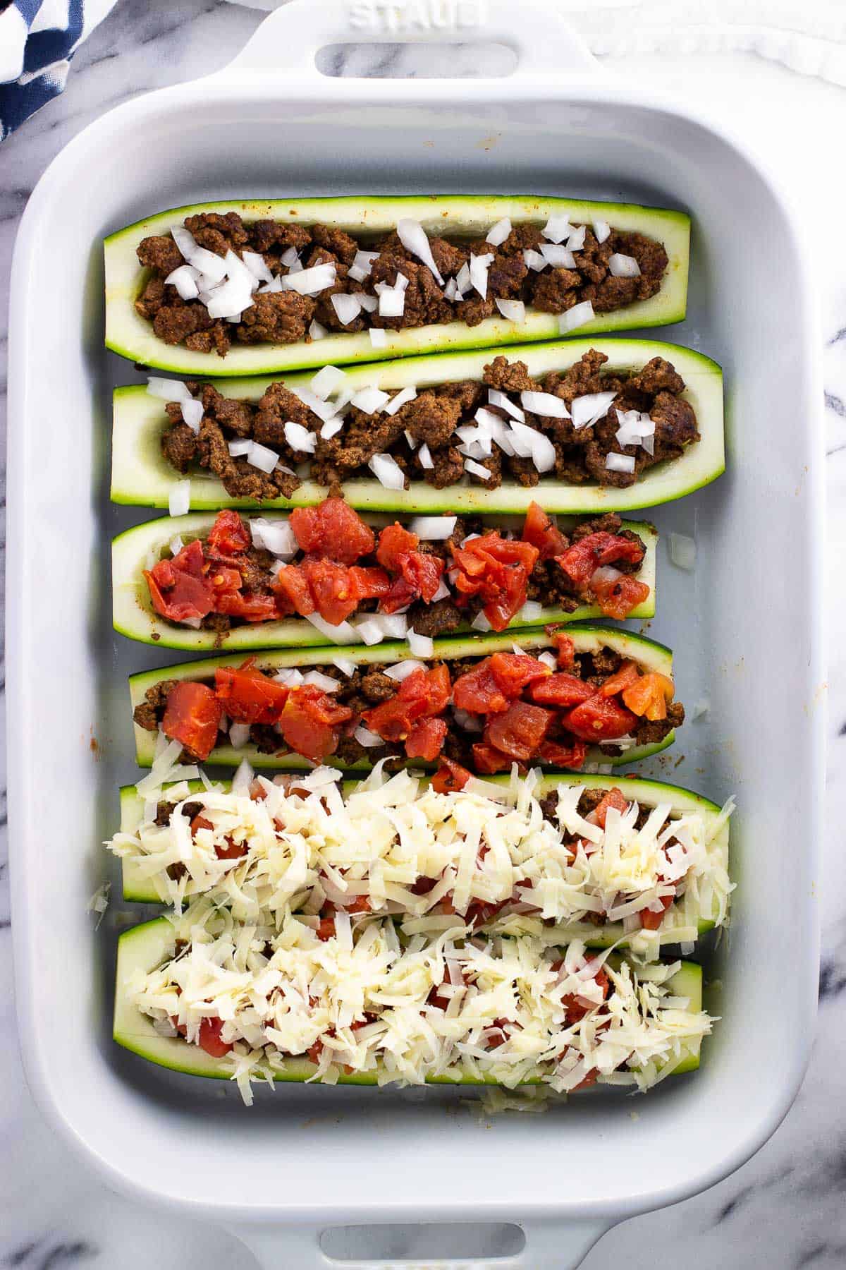 Six zucchini boats in a baking dish in the process of having toppings added.