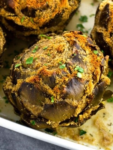 A close-up of a stuffed artichoke in a pan of cooking liquid.