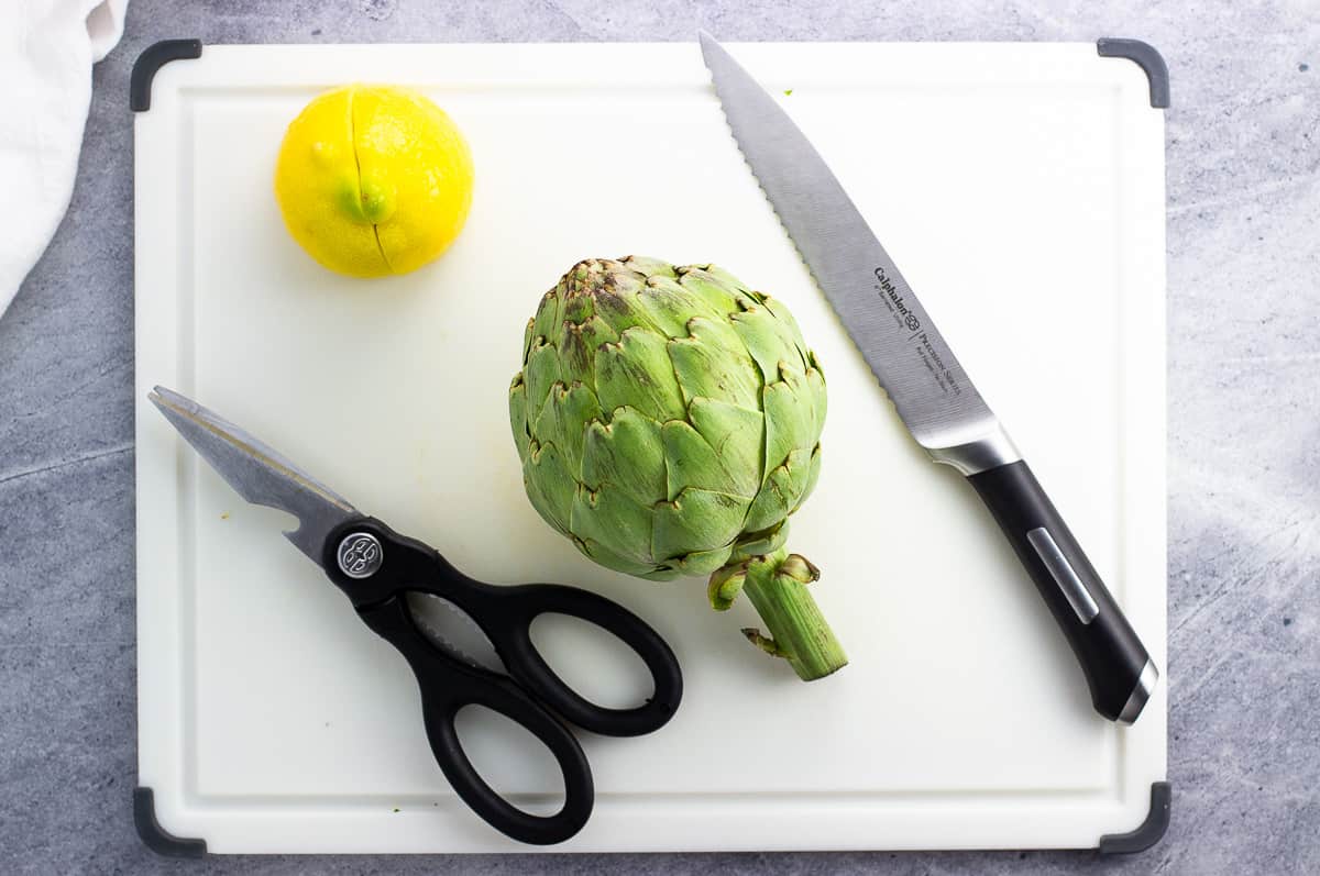 Half a lemon, a whole artichoke, kitchen shears, and a serrated knife on a cutting board.